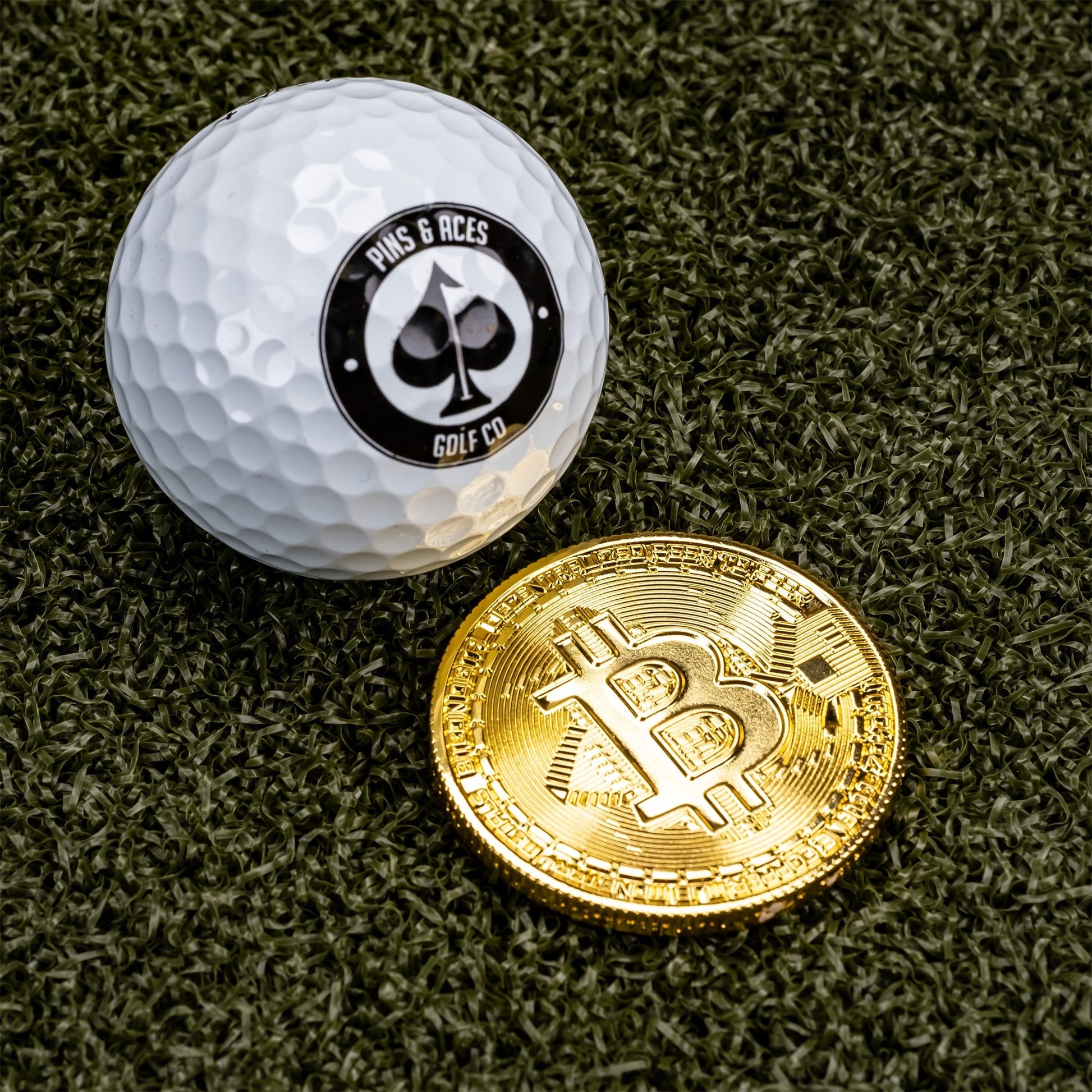 Bitcoin ball marker beside a golf ball featuring the Pins and Aces logo, set on a green turf background.