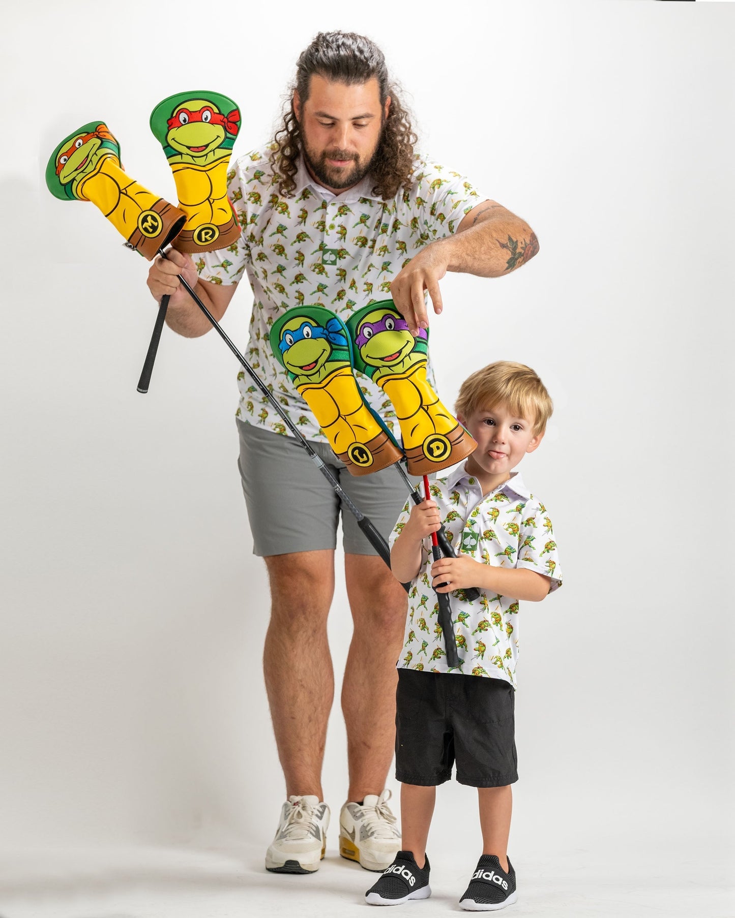 A child and adult hold colorful TMNT-themed golf headcovers, both wearing matching playful shirts, ready for a fun day on the course.