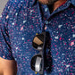 Men's blue splatter golf shirt featuring vibrant paint splashes, paired with stylish sunglasses hanging from the collar.