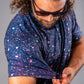 A golfer adjusts the sleeve of a blue splatter polo shirt, showcasing its vibrant, colorful design against a gray background.