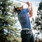 A golfer swings a driver while wearing a blue splatter-patterned polo shirt and black shorts on a sunny day.