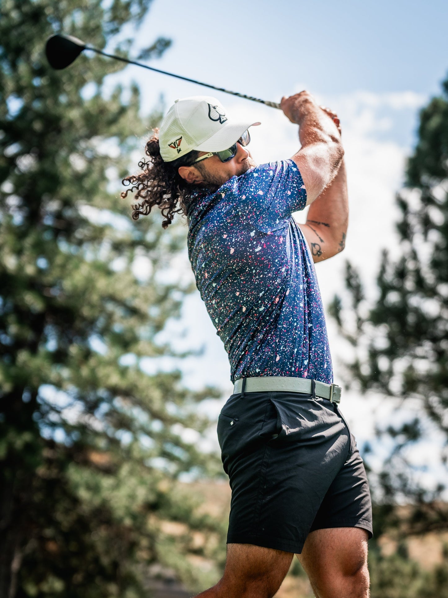 A golfer swings a driver while wearing a blue splatter-patterned polo shirt and black shorts on a sunny day.