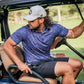 A golfer wearing a vibrant lavender floral polo shirt and a cap sits in a golf cart on the course.