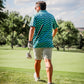 Man walking on a golf course wearing a wavy checkered blue and green polo shirt and shorts, holding a golf club.