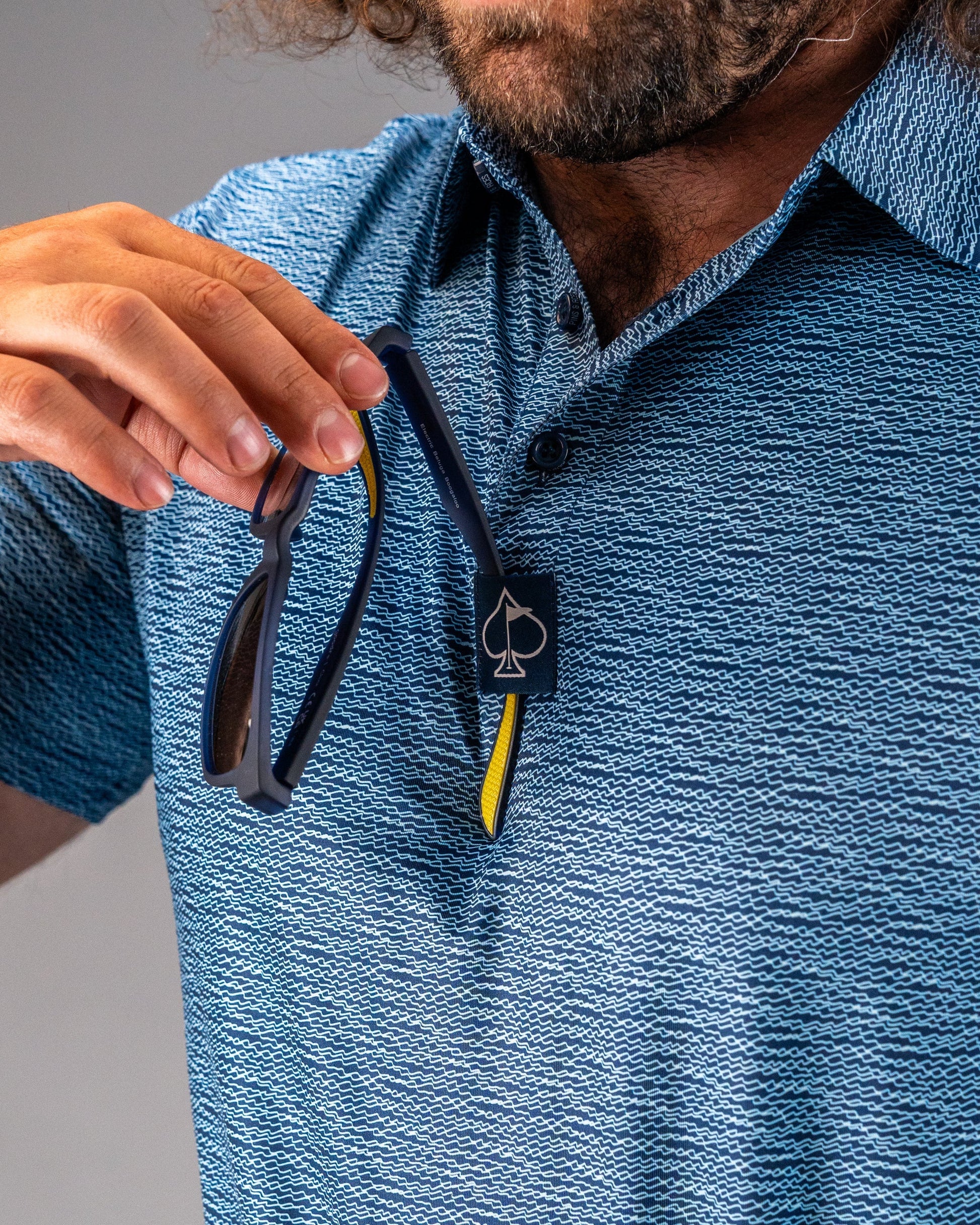 A golfer holds sunglasses while wearing a blue patterned polo shirt with a logo on the collar, showcasing stylish golf apparel.