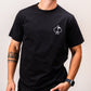 Man wearing a black t-shirt featuring a white spade symbol on the chest, styled with a casual wristwatch and displaying a tattoo on his left arm.