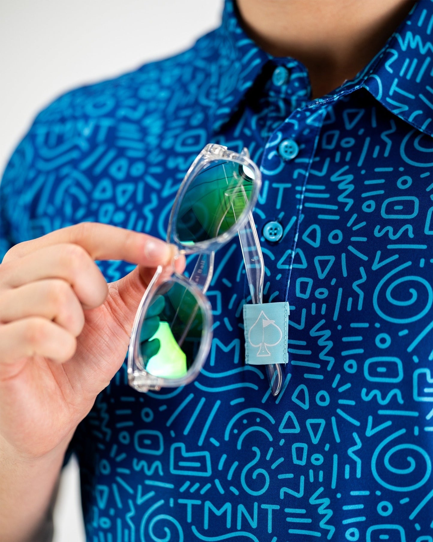 A person holds sunglasses while wearing a blue TMNT-themed shirt featuring a doodle pattern.