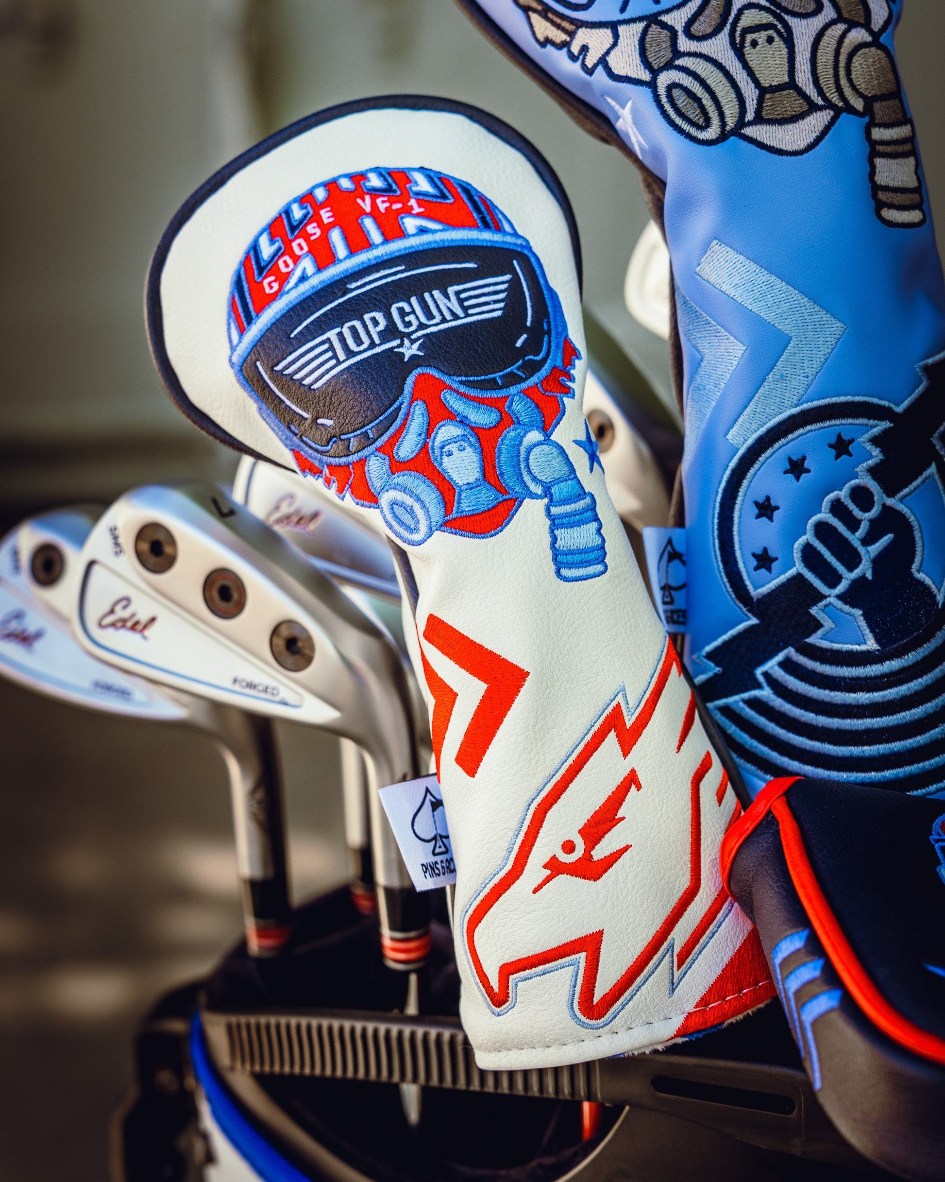 Top Gun Goose themed golf club headcover with embroidered logo and colorful design, placed among other golf club headcovers.